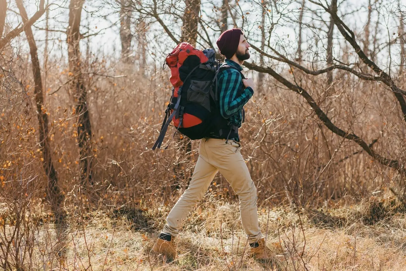 Attraktiver Hipster-Mann reist mit Rucksack in einem herbstlichen Wald und trägt ein kariertes Hemd sowie einen Hut.